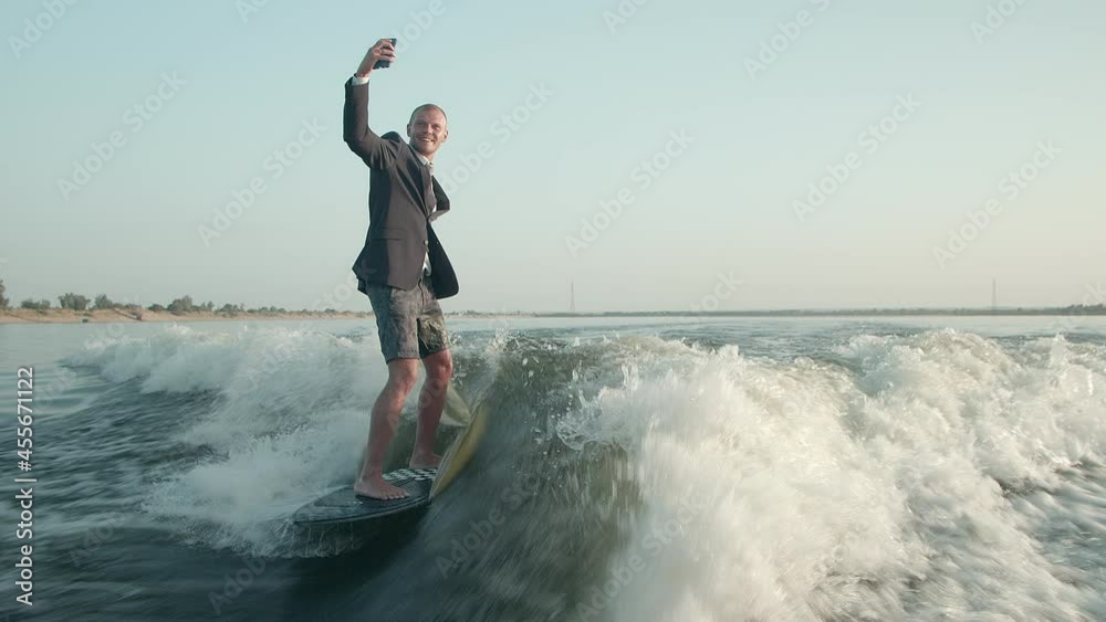 A surfer jumping on a wakeboard in a suit with a phone in his hands takes pictures of himself. A surfer takes a selfie and rides a wakeboard. A surfer jumping on a wakeboard in a suit with a phone in his hands takes pictures of himself. A surfer takes a selfie and rides a wakeboard.