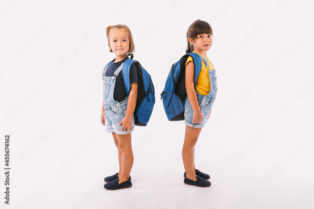 Obraz premium Two little girls, one with blonde hair and the other with black hair, dressed in denim blue overalls, with a backpack, ready for back to school, sideways, on white background.