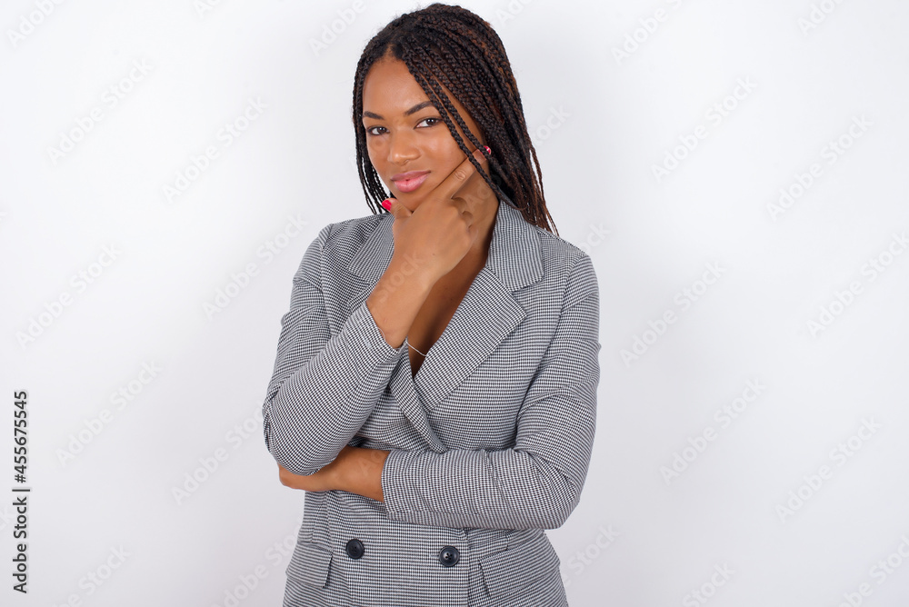 Thoughtful smiling Young african american business woman with braids over white wall keeps hand under chin, looks directly at camera, listens something with interest. Youth concept. Thoughtful smiling Young african american business woman with braids over white wall keeps hand under chin, looks directly at camera, listens something with interest. Youth concept.