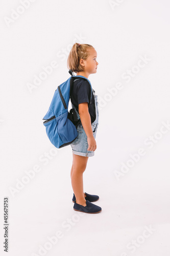 Little girl with blond hair dressed in a denim overalls and a blue t-shirt, with a backpack ready for going back to school, on her side, on white background