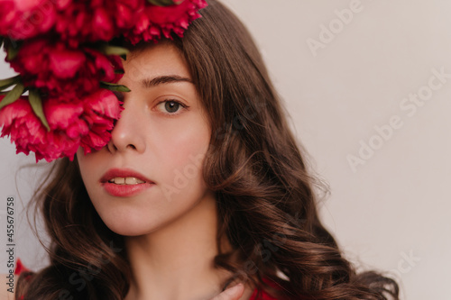 Full close up photography of brown hair grey eyes lady watching directly into the camera. Wonderfull peonies bouquet, summer cherry lipstick. High quality photo above white wall with soft light