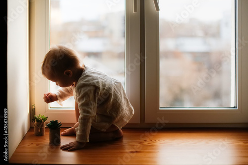 A cute toddler child on the windowsill by the window playing with plant, children's safety in apartments, a cozy photo in a Scandinavian interior