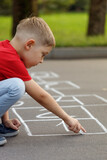 Cute little boy drawing hopscotch with white chalk on playground.