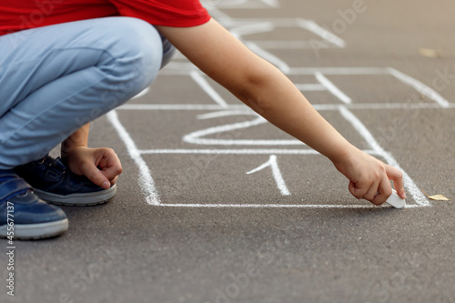 Little boy in red t-shirt drawing hopscotch with chalk on playground. Close up photo with blur background.