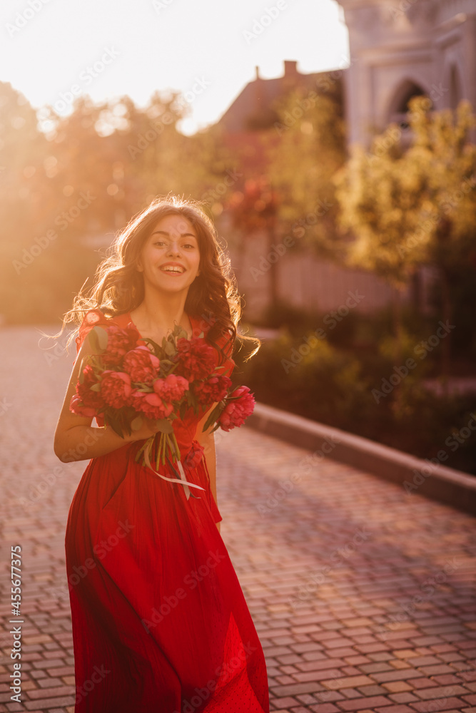 Magical sunny light outside, medium full shot portrait of attractive lady with bouquet. Dark curly hair, ruby summer dress and smiles with red lipstick looks romantically with the cherry peony 