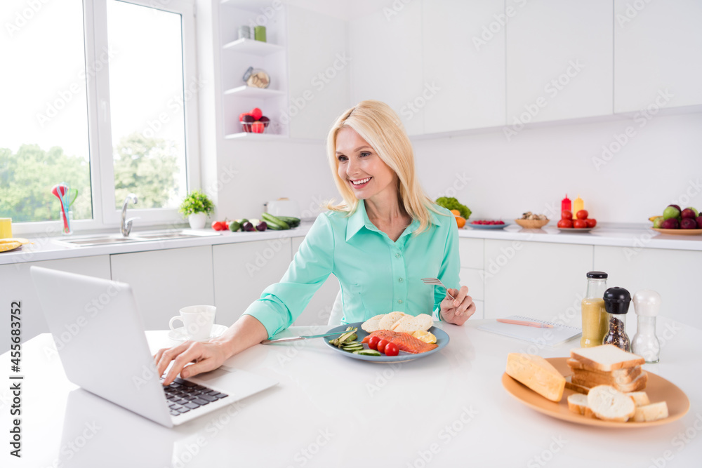 Photo of charming blonde lady sit table have lunch browse netbook wear green shirt kitchen room indoors