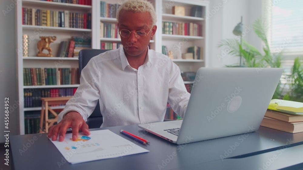 Serious African American businessman prepares data for a marketing report by comparing graphs on paper and laptop. Millennial man in glasses and shirt working in the office of a modern business center