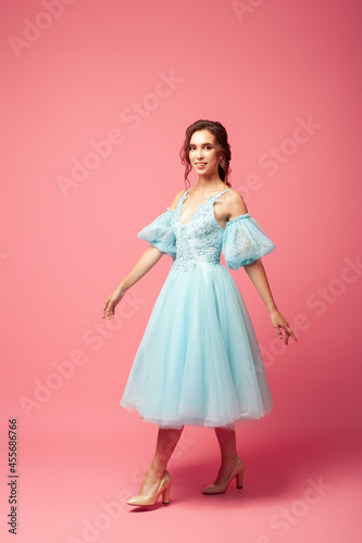 full-length portrait in motion, walking, smiling young woman in blue evening dress with full skirt and lantern sleeves, isolated on pink background. 