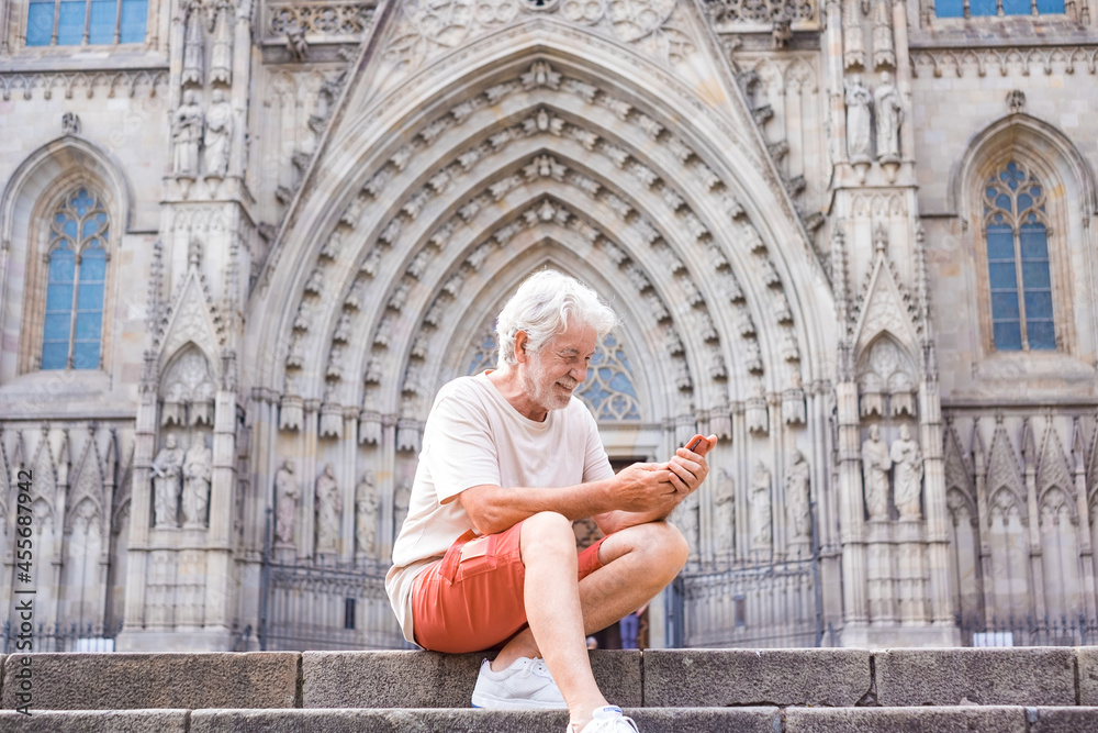 Smiling senior white-haired traveler enjoying visit to cathedral of Barcelona, sitting on staircase holding mobile phone. Retired man enjoying free time and journey