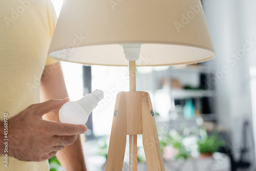 Cropped view of man holding energy saving light bulb near floor lamp at home