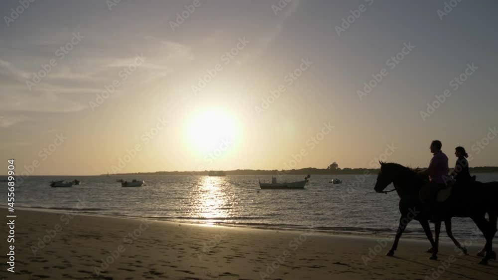 Backlighting of a couple strolling on horseback at sunset on the shore of Bajo de Guía beach in Sanlúcar de Barrameda Cádiz with fishermen's boats in the background. Backlighting of a couple strolling on horseback at sunset on the shore of Bajo de Guía beach in Sanlúcar de Barrameda Cádiz with fishermen's boats in the background.
