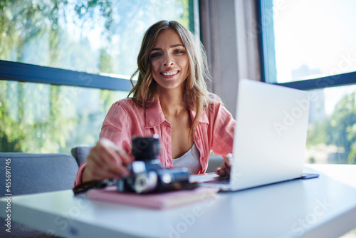 Portrait of cheerful professional photographer enjoying time for doing journalistic distance job in cafe interior, happy Caucaisan freelancer smiling at camera at desktop with netbook device