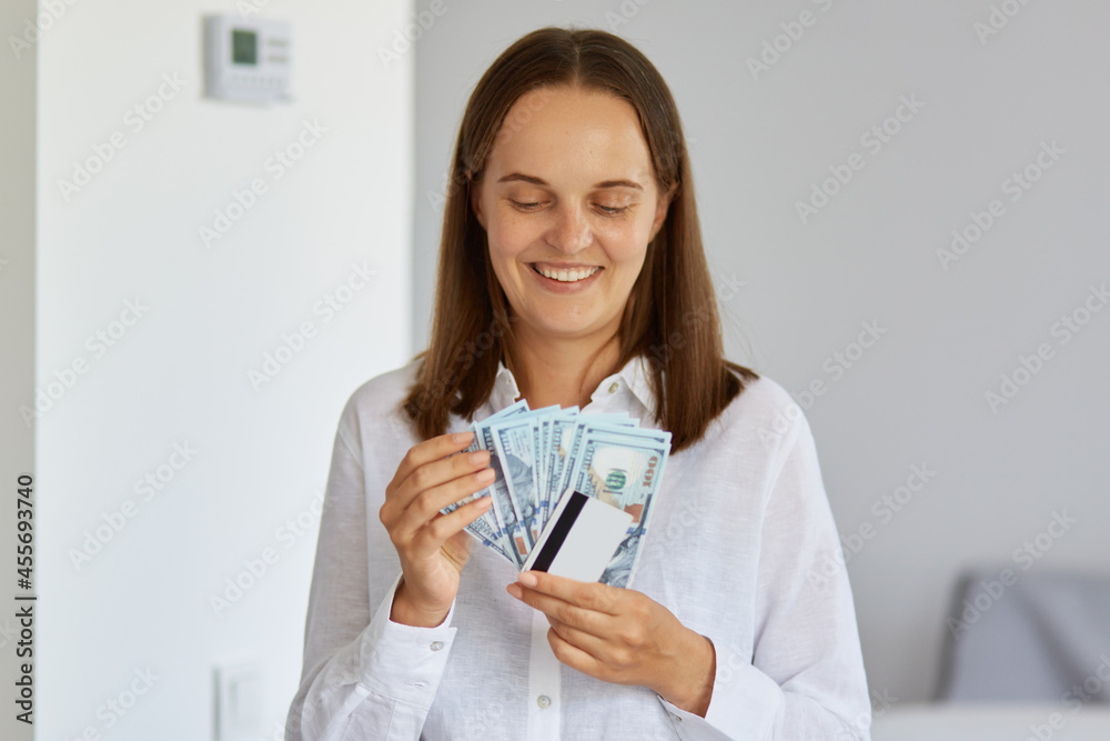 Indoor shot of positive young woman standing in light room at home, wearing white shirt, holding money and credit card, looking at banknotes with toothy smile. Indoor shot of positive young woman standing in light room at home, wearing white shirt, holding money and credit card, looking at banknotes with toothy smile.