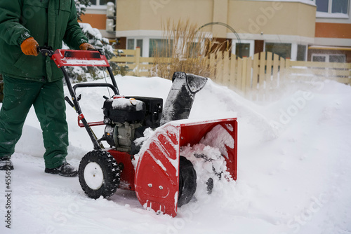 A worker in green overalls removes snow with a red snowblower against the background of a spruce, a snowdrift and a house.