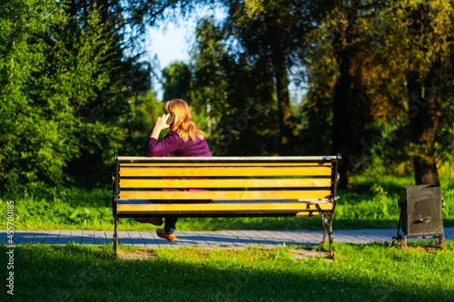 Defocus caucasian blond woman talking, speaking on the phone outside, outdoor. 45s years old woman in purple blouse in park on bench. Adult women using telephone. Back view. Copy space. Out of focus