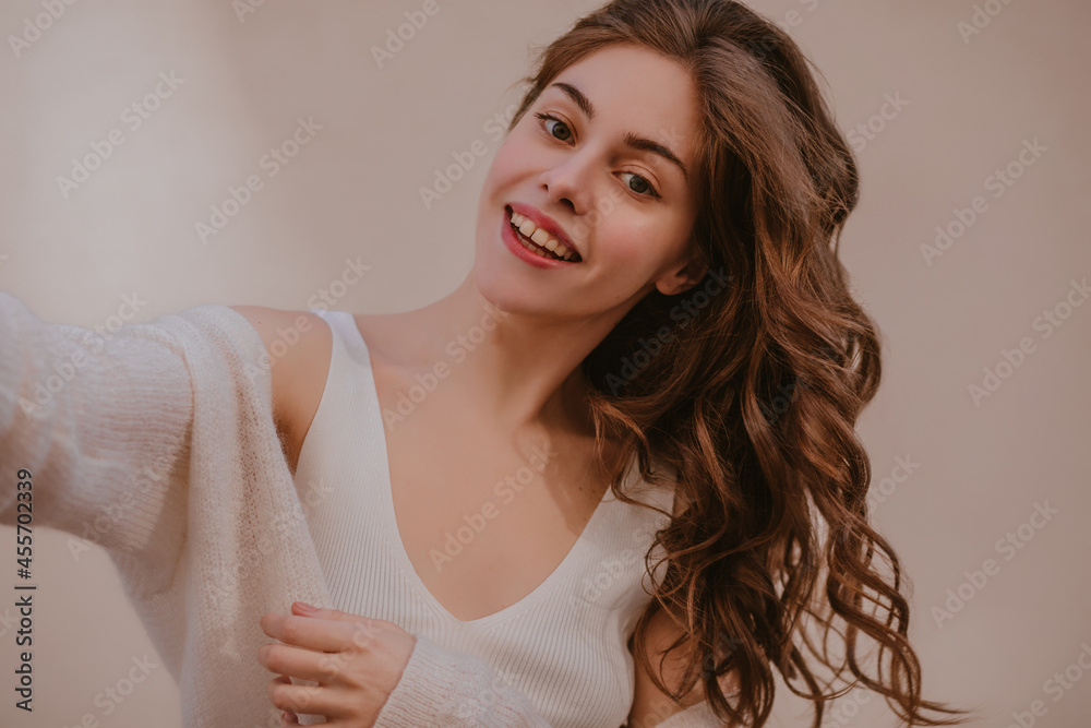Close-up of fair-skinned lady makes selfie smiling with teeth on light background. Long blond hair scatters on one side. A girl in white T-shirt and warm sweater on top of her shoulders. Close-up of fair-skinned lady makes selfie smiling with teeth on light background. Long blond hair scatters on one side. A girl in white T-shirt and warm sweater on top of her shoulders.