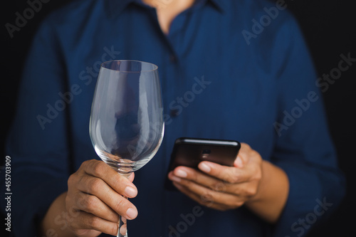 Midsection of a man holding an empty glass of wine and using a smartphone while standing with a black background