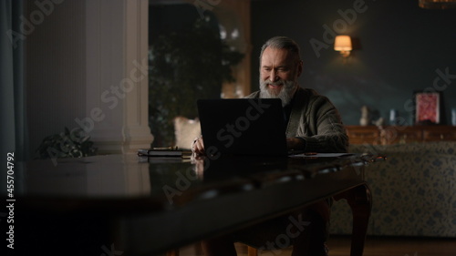 Joyful old man making video call on computer in luxury cabinet. Happy senior man
