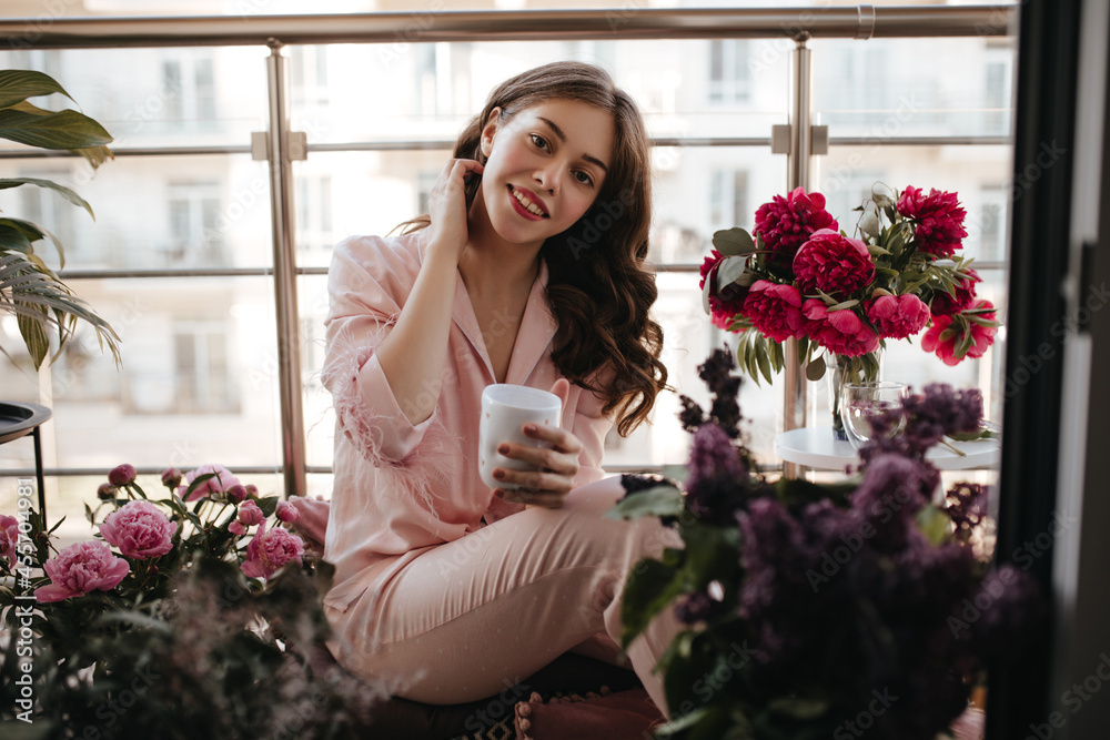 Joyful belarusian lady sitting on the balcony floor during saturday morning. Drinking tea in her lovely soft pink pajamas, brunette smelling peonies bouquets all around and touching her neck