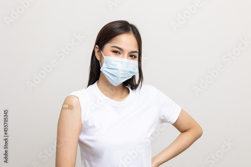 Young beautiful asian woman wearing mask and getting a vaccine protection the coronavirus. Happy female showing arm with bandage after receiving vaccination on isolated white background.