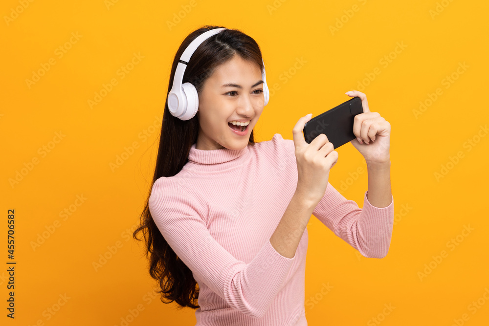 Beautiful young asian women play mobile game and put on wireless headphone standing on isolated yellow background. Playing game on smartphone winning victory moment. Very enjoy and fun relax time Beautiful young asian women play mobile game and put on wireless headphone standing on isolated yellow background. Playing game on smartphone winning victory moment. Very enjoy and fun relax time