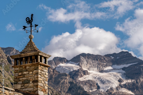 A rooster-shaped weather vane atop a chimney in Lillaz, Aosta Valley, Italy, with the Gran Paradiso glacier in the background