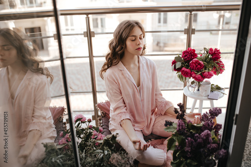 Young pleasent moldavian female meditating among fresh flowers with closed eyes. Brunette girl with natural face in wonderful pink chiffon homewear relaxing on her cozy balcony with beautiful sunlight photo