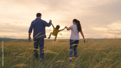 Mom, daughter, dad are playing on field in sunshine, happy child is holding hands of his parents and jumping. Family weekend. Happy family runs together holding hands in spring, summer. Teamwork.