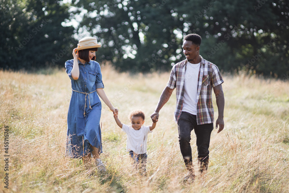 Beautiful and happy multiracial family walking on wild field during warm summer day. Young mother and father holding hands with cute little son. Time spending on fresh air. Beautiful and happy multiracial family walking on wild field during warm summer day. Young mother and father holding hands with cute little son. Time spending on fresh air.