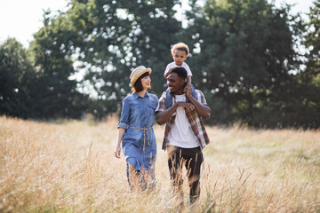 African american man carrying his little son on shoulders while walking with beautiful caucasian wife outdoors. Portrait of happy multiracial family. Love concept.
