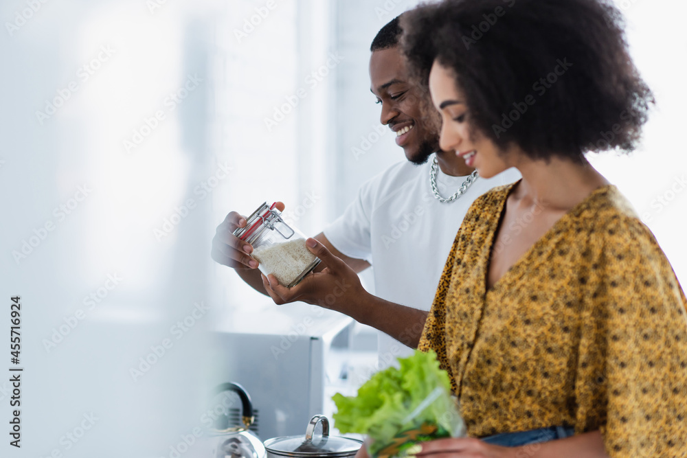 Side view of african american man holding jar with rice near blurred girlfriend in kitchen. Side view of african american man holding jar with rice near blurred girlfriend in kitchen.