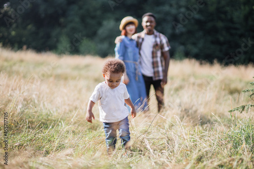 Pretty african boy running and playing among high grass while his multiracial parents standing in hugs on blur background. Concept of family, happiness and lifestyles.