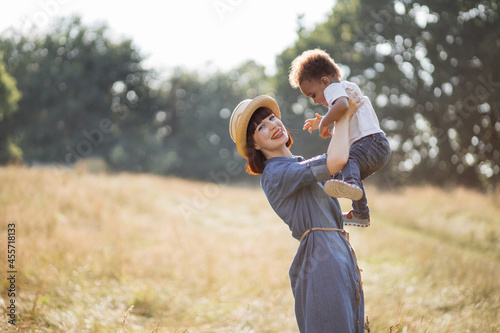 Smiling caucasian woman lifting her lovely black child while taking fun on summer field. Multiracial family outdoors. Happiness and love concept.