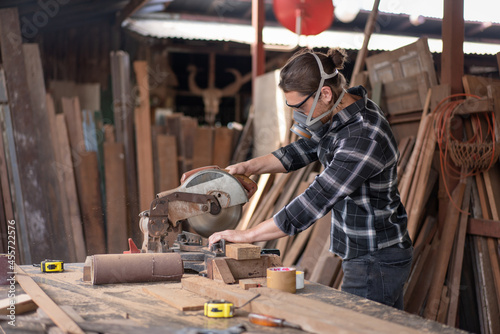 Male carpenter wears a mask to prevent wood chips from entering his face while using a circular saw machine to cut woodwork in a carpentry workshop.