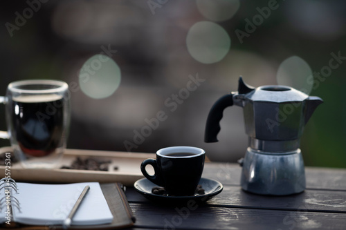 Photo of coffee cup putting on a wooden table surrounded by Moka pot and notebook over a nature outdoors as a background.