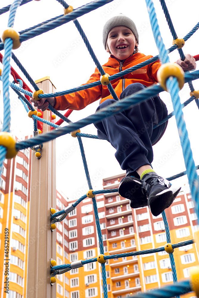 Funny caucasian blond boy in an orange jacket plays on the sports playground.