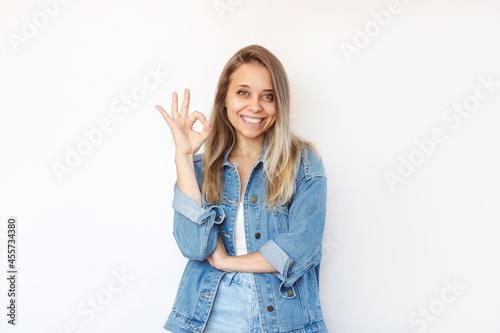 A young caucasian smiling happy pretty blonde woman in a denim jacket and jeans showing okay gesture with her hand isolated on a white background. Positive human emotion. OK approval sign