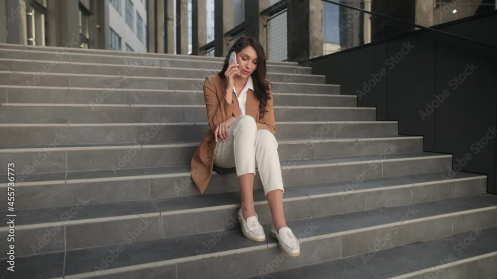 Smiling young business woman talking on phone sitting on office stairs. Portrait of friendly lady. High quality 4k footage Smiling young business woman talking on phone sitting on office stairs. Portrait of friendly lady. High quality 4k footage