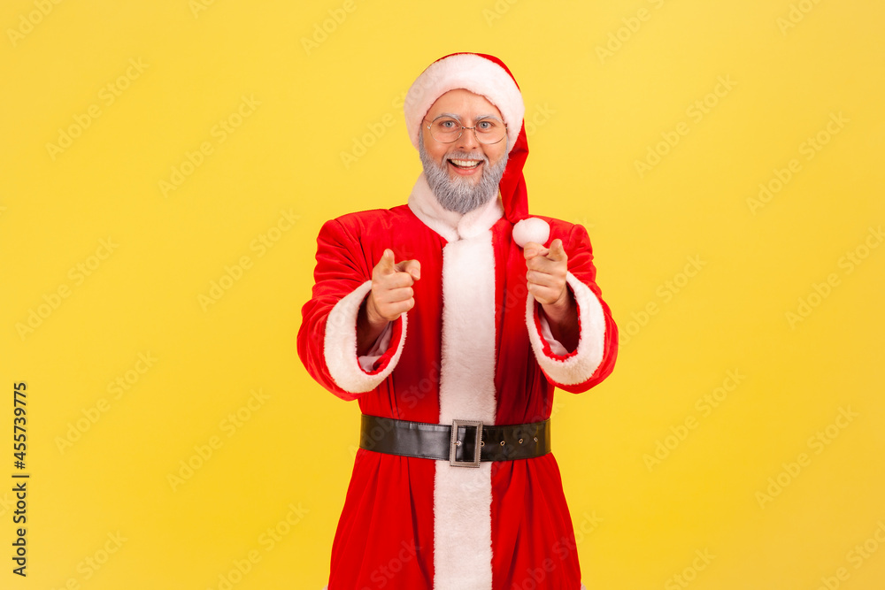 Pleasant looking positive elderly man with gray beard wearing santa claus costume standing pointing to camera with smile, choosing you. Indoor studio shot isolated on yellow background. Pleasant looking positive elderly man with gray beard wearing santa claus costume standing pointing to camera with smile, choosing you. Indoor studio shot isolated on yellow background.