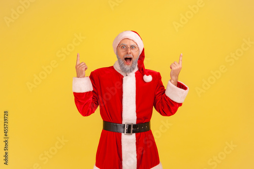 Excited screaming elderly man with gray beard in santa claus costume pointing up with both hands  keeps mouth open  presenting copy space above head. Indoor studio shot isolated on yellow background.