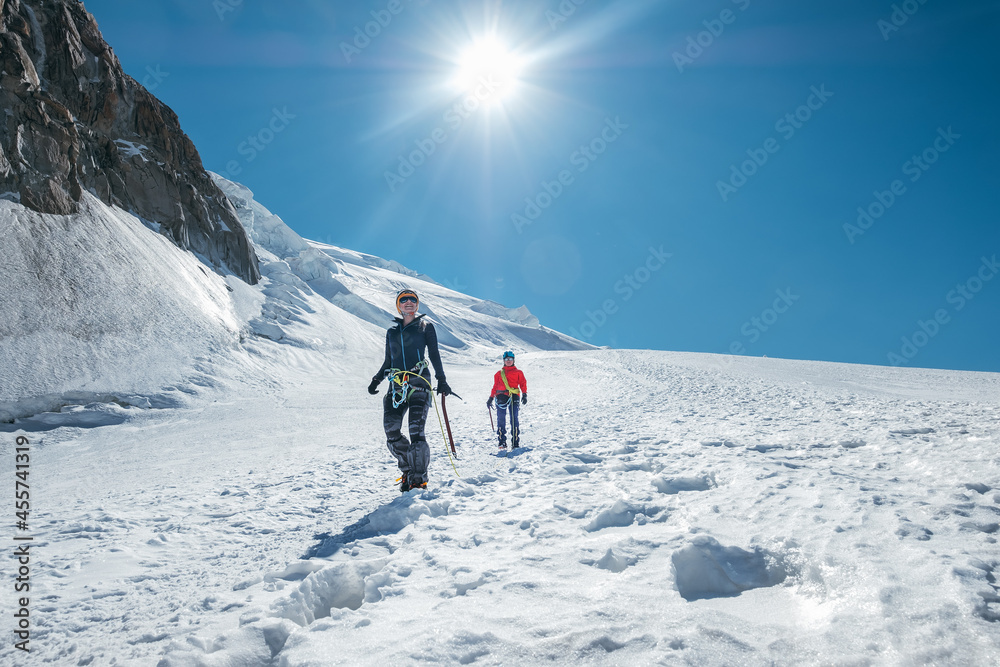 Two laughing young women Rope team descending Mont blanc du Tacul summit 4248m dressed mountaineering clothes with ice axes walking by snowy slopes. People extreme activities sporty concept image.