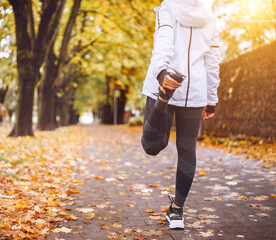 Fit athletic woman doing stretching before jogging in the autumnal city park. Young fitness female runner stretching legs while warming up. Active running people concept image.