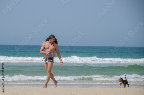 man gay walking in beach clothes, hat and sunglasses, on the sand with his little black dog, in the background the blue sea and its clear on a summer day 