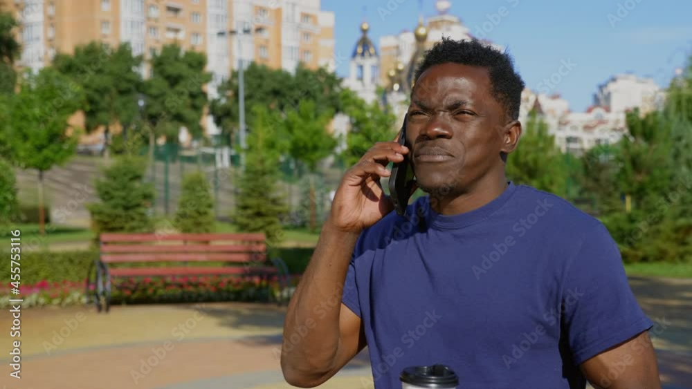 Afro-American man standing in park talking on cell phone and looks around. African adult male ethnic black wears blue t-shirt uses smartphone in summer Afro-American man standing in park talking on cell phone and looks around. African adult male ethnic black wears blue t-shirt uses smartphone in summer