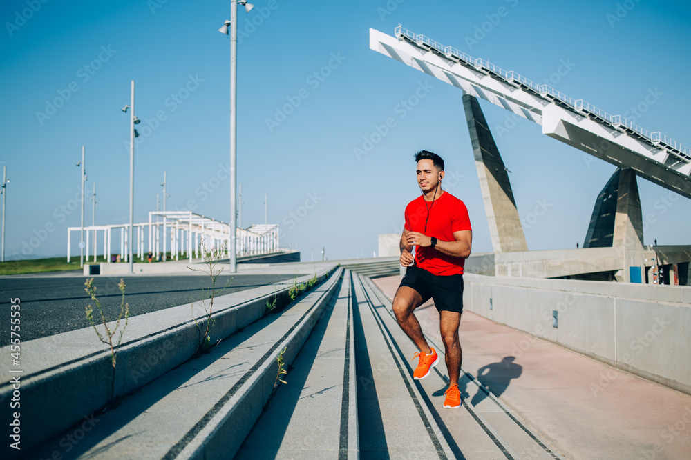 Motivated male jogger dressed in active wear running and listening music player in earphones enjoying sportive lifestyle, good looking man in tracksuit have energetic cardio training at urbanity