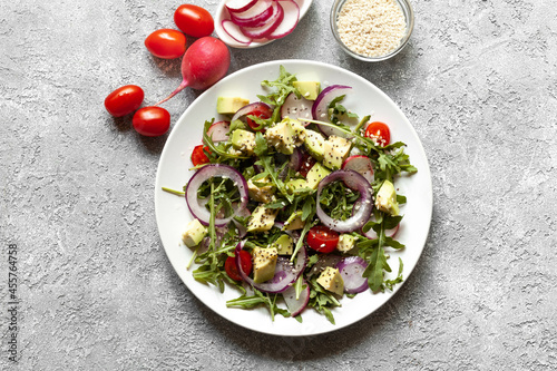 Fresh vegan salad with arugula, tomatoes, avocado, radish and red onion on grey texture