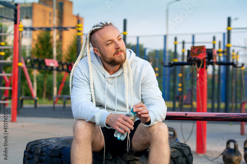 A man with an interesting braid hairstyle drinks water from a bottle after a workout on an outdoor fitness site.