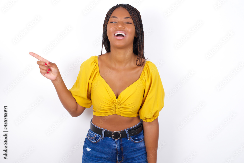 Young african american woman with braids over white wall laughs happily points away on blank space demonstrates shopping discount offer, excited by good news or unexpected sale. Young african american woman with braids over white wall laughs happily points away on blank space demonstrates shopping discount offer, excited by good news or unexpected sale.