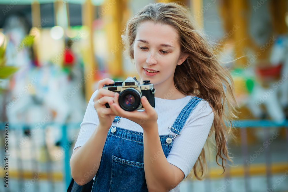 A young woman tourist in a denim sundress stands with cameras in an amusement park.
