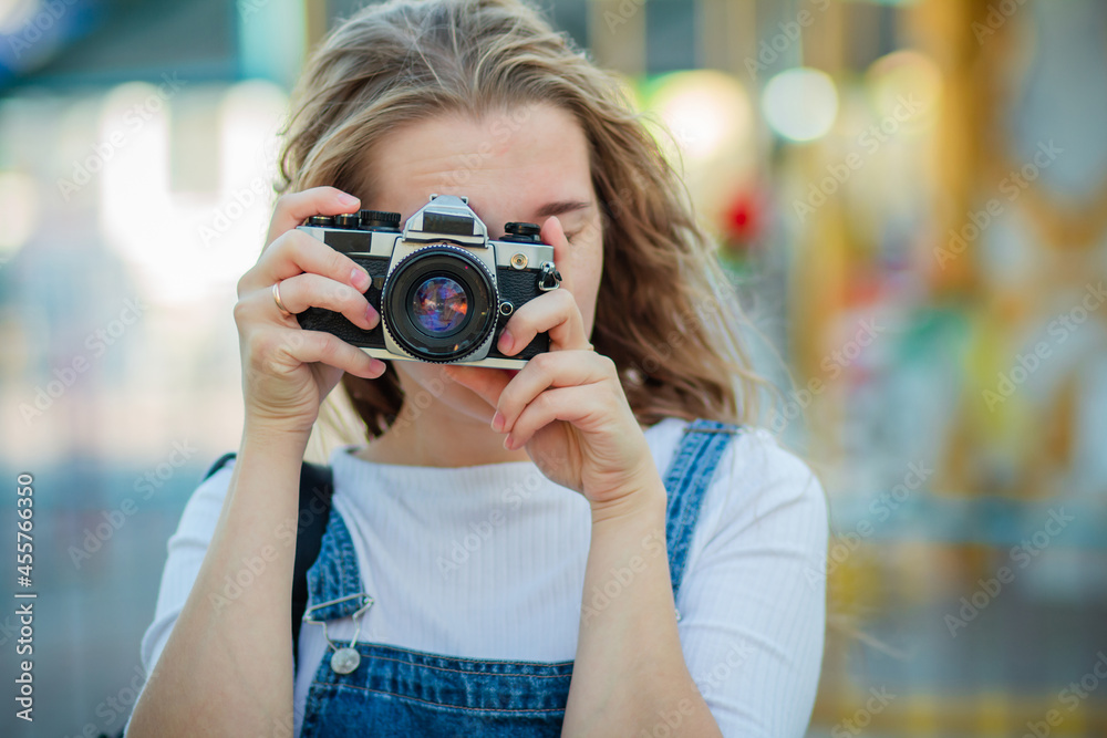 A young woman tourist in a denim sundress stands with cameras in an amusement park.